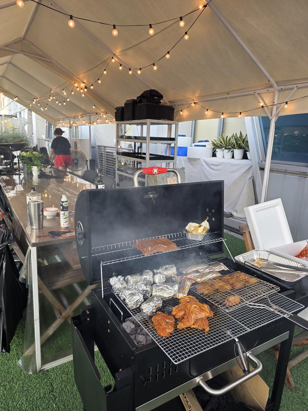 A charcoal grill loaded with chicken, foil-wrapped parcels and skewers under the fairy-lit farm pavilion - farm-to-table cooking at the Harvest & Cook Experience
