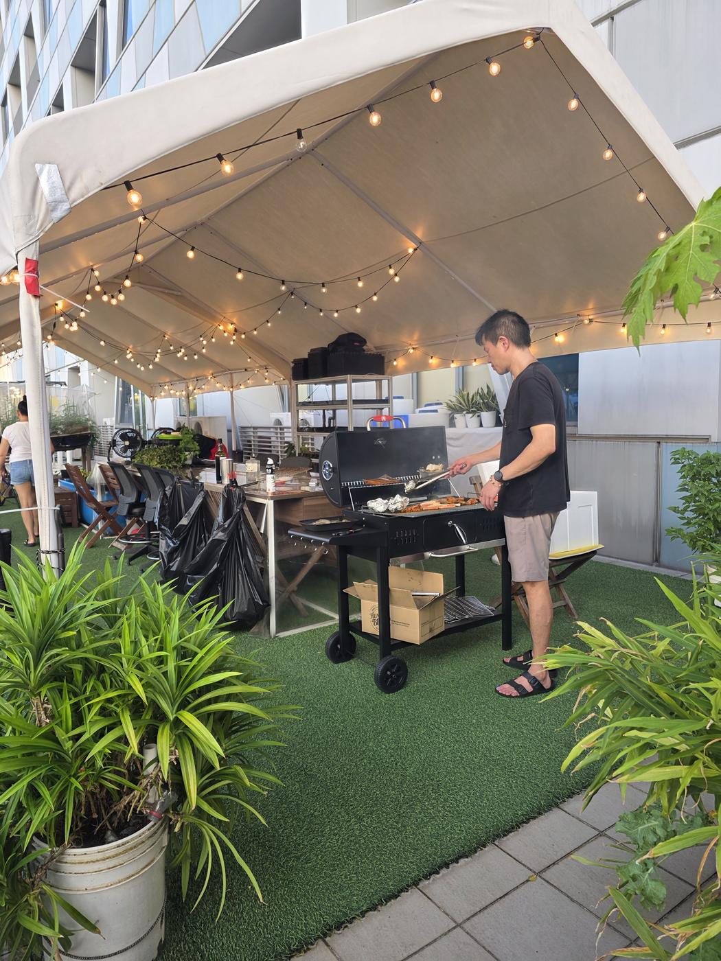 A guest grilling at the charcoal BBQ under the string-lit sheltered pavilion, surrounded by greenery